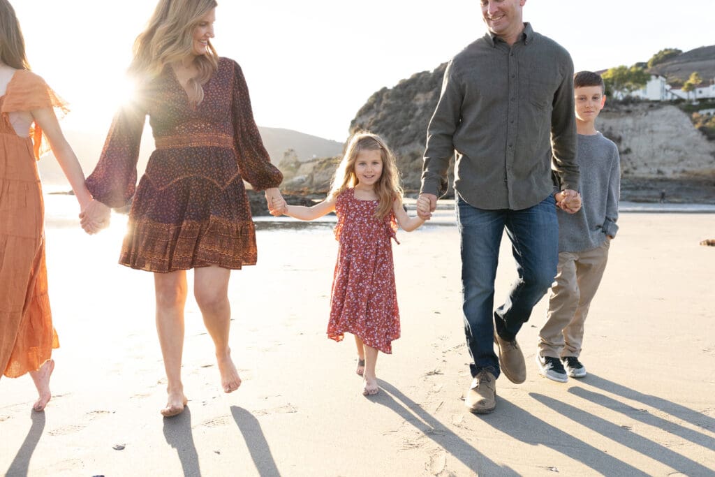Family walking barefoot along the beach at sunset, holding hands with sunlight streaming through, capturing a relaxed and joyful moment.