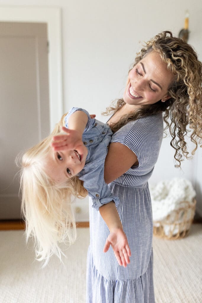 Mother laughing and playfully holding her daughter upside down in a cozy home setting, a joyful moment that highlights the value of choosing the right photographer.