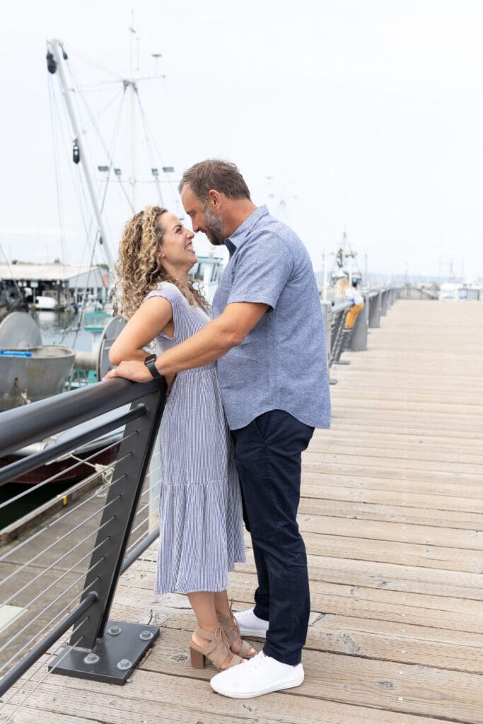 Couple smiling and embracing on a dock beside fishing boats, sharing a quiet, joyful moment together on a cloudy day.