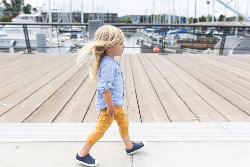 Young child with long blonde hair walking confidently along a dock near boats, wearing a blue shirt and mustard yellow pants on a breezy day.