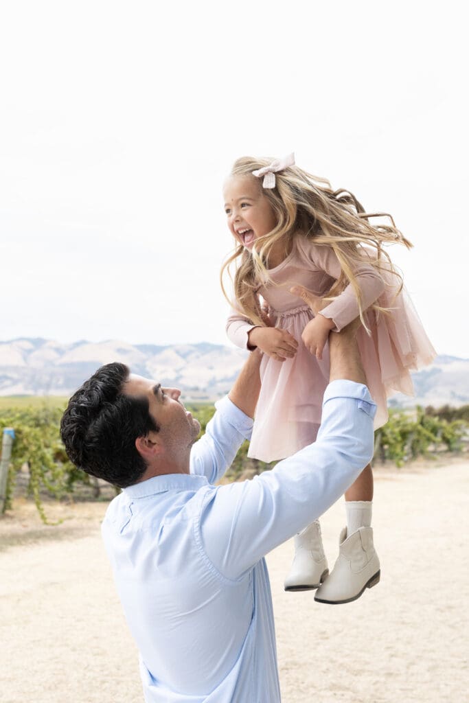 Father lifting his smiling daughter into the air in a vineyard setting, capturing the playful joy families want when choosing the right photographer.