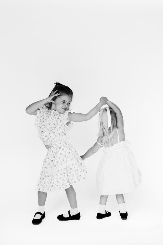 Two young girls twirling joyfully in dresses, capturing playful, candid energy in family photos at home.