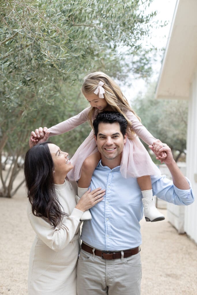 Father smiling with daughter on his shoulders while mother looks up at them, sharing a joyful moment outdoors with soft natural light.