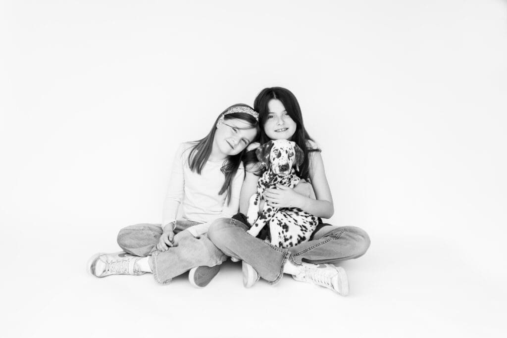Two girls sitting and cuddling a Dalmatian puppy during a candid kids photo session in studio, black and white.
