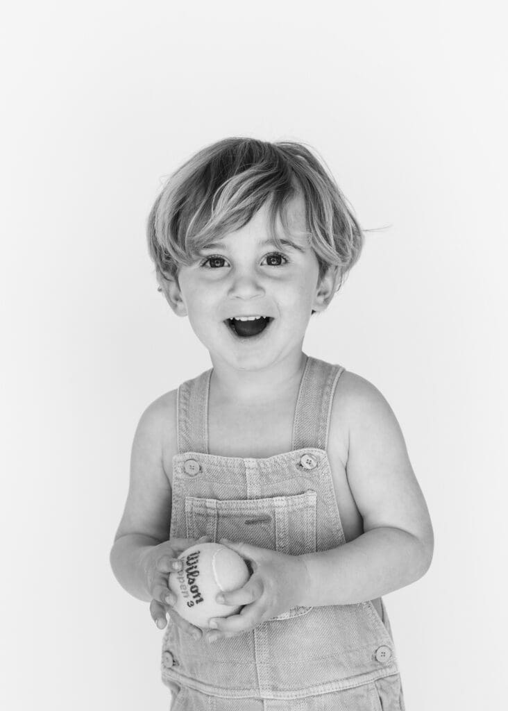 Toddler holding a tennis ball and smiling during a  photo session in studio, black and white portrait.