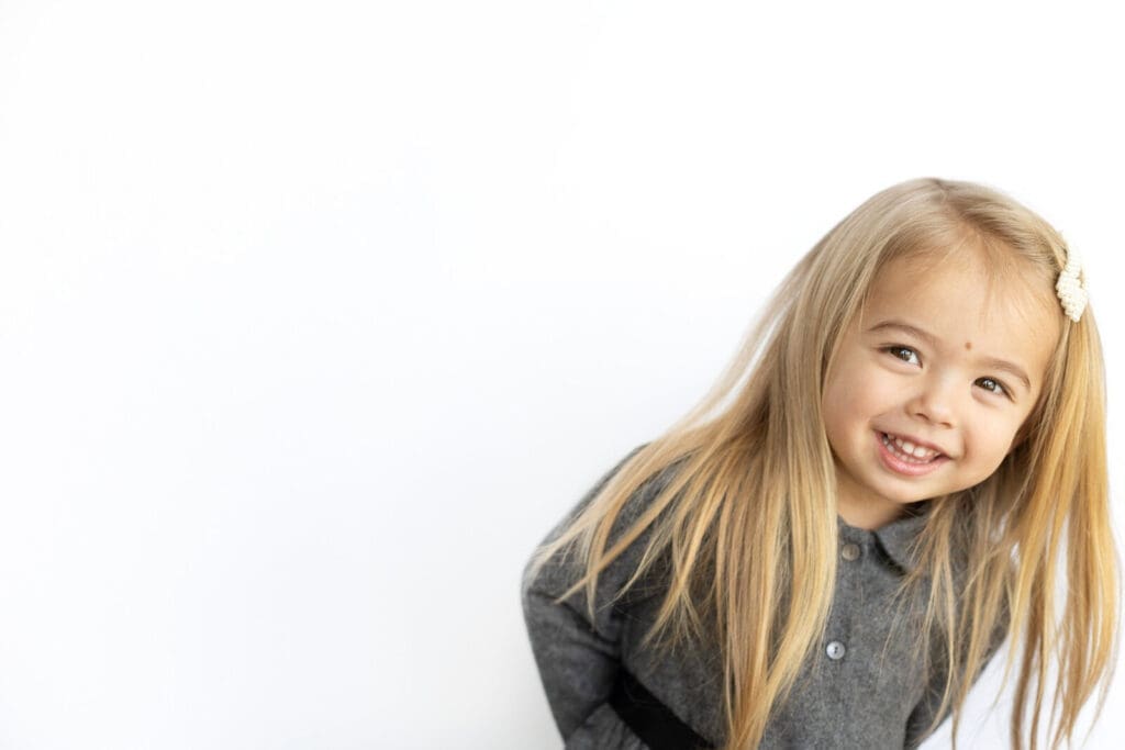 Smiling young girl with long blonde hair leaning playfully during preschool photos against a white background.
