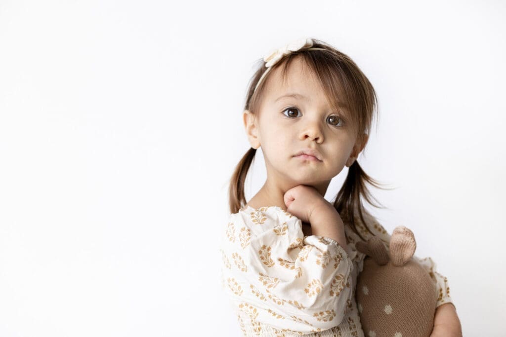 Young girl with pigtails and floral dress holding a stuffed toy, looking softly at the camera.