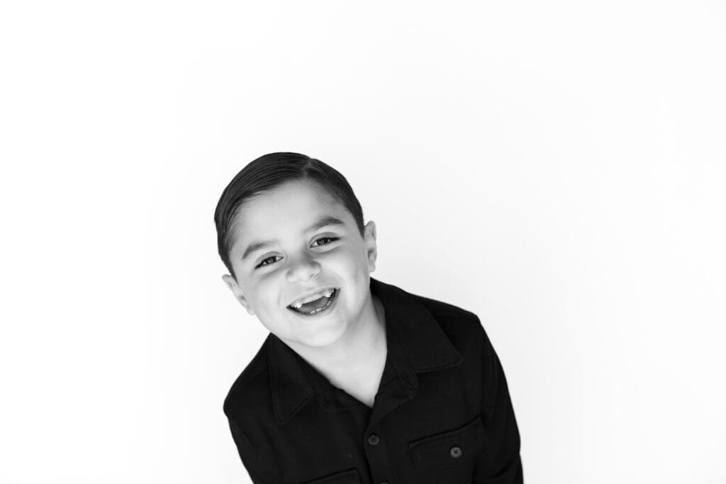 Laughing young boy in dark shirt leaning forward during preschool photos against a bright white background.