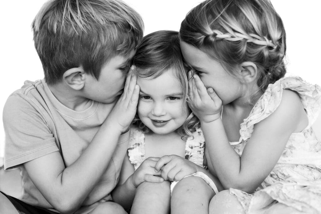 Two kids whisper secrets into a smiling girl’s ears, creating a sweet and playful black-and-white portrait.