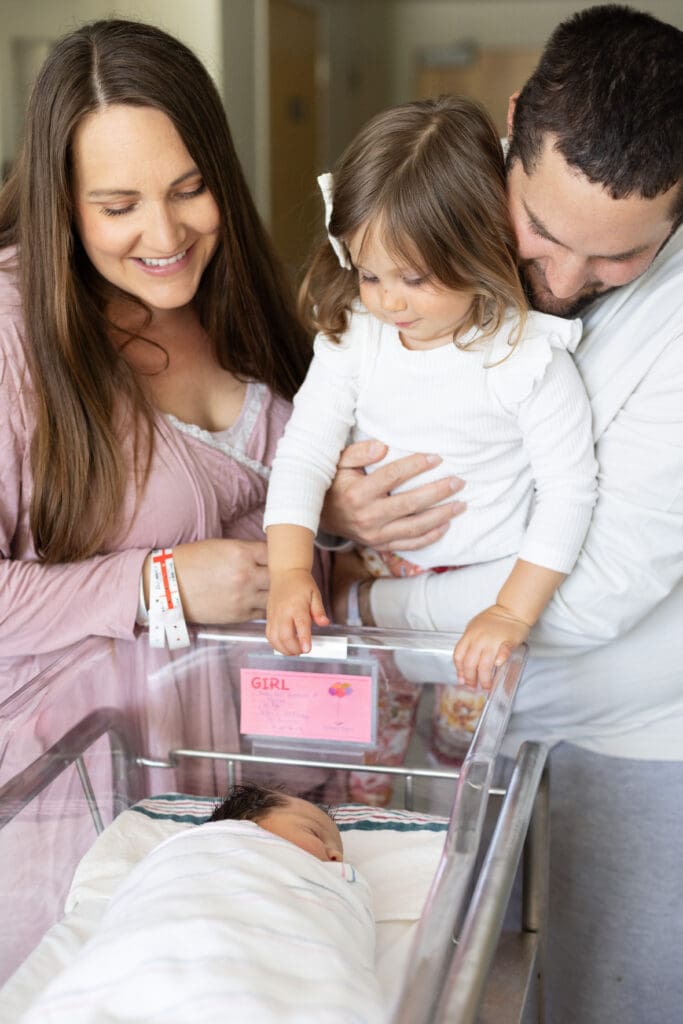 Parents smile while holding toddler who looks into newborn’s hospital bassinet during family’s first moments together.