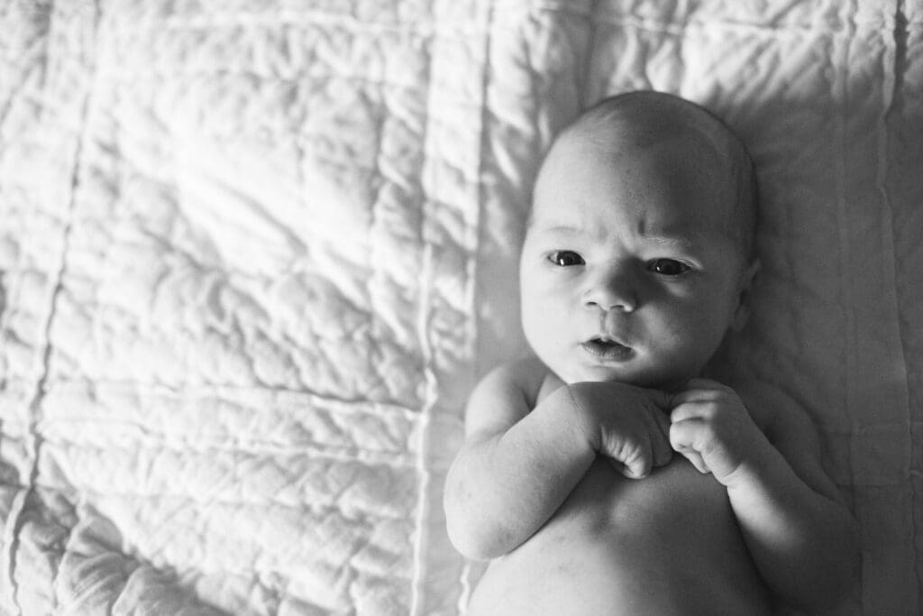 Black and white photo of newborn baby lying on quilted blanket with hands near face.