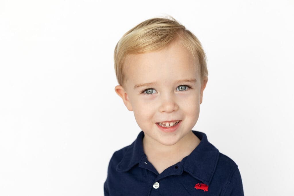 Smiling young boy with blonde hair and blue eyes wearing a navy polo shirt, standing against a bright white background during his preschool photos.