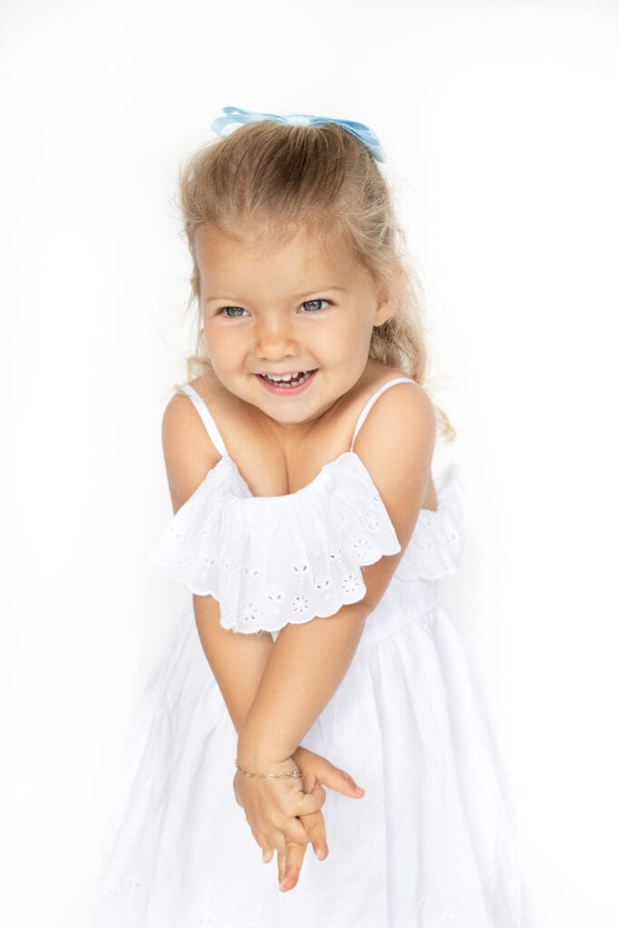 Smiling little girl in a white dress with eyelet ruffles, hands clasped playfully, wearing a blue hair bow.