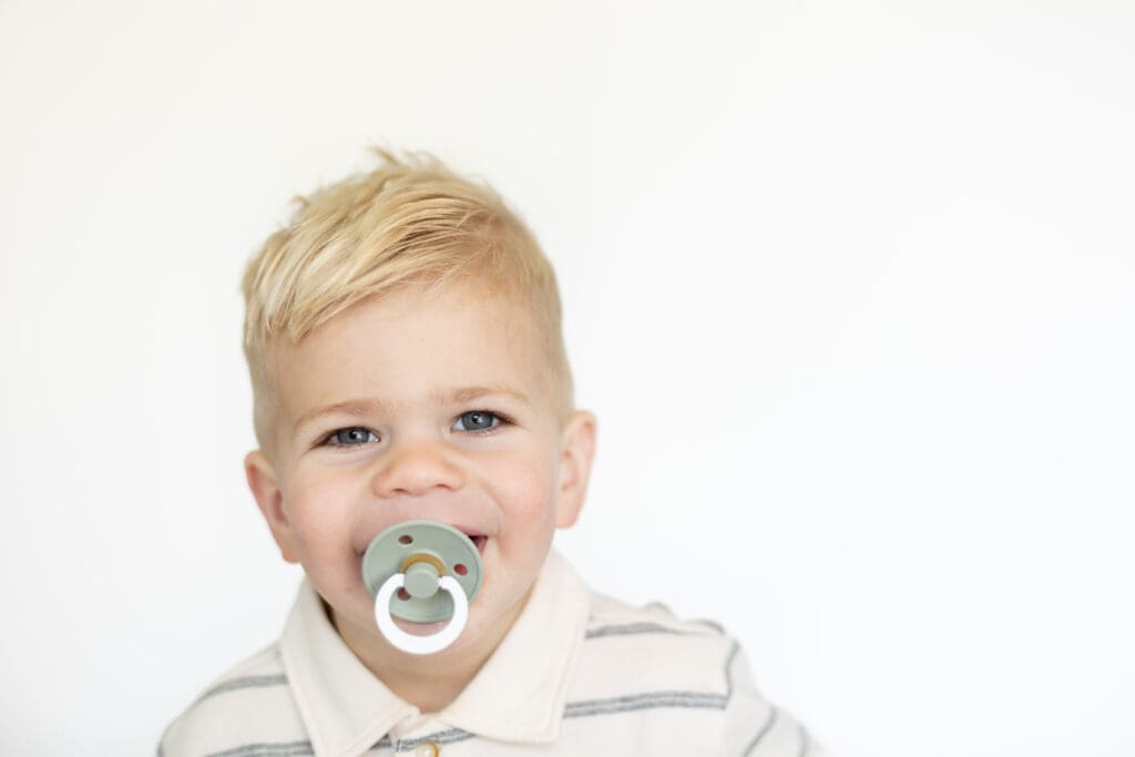 Blonde toddler with a pacifier in his mouth, grinning in a light striped shirt.