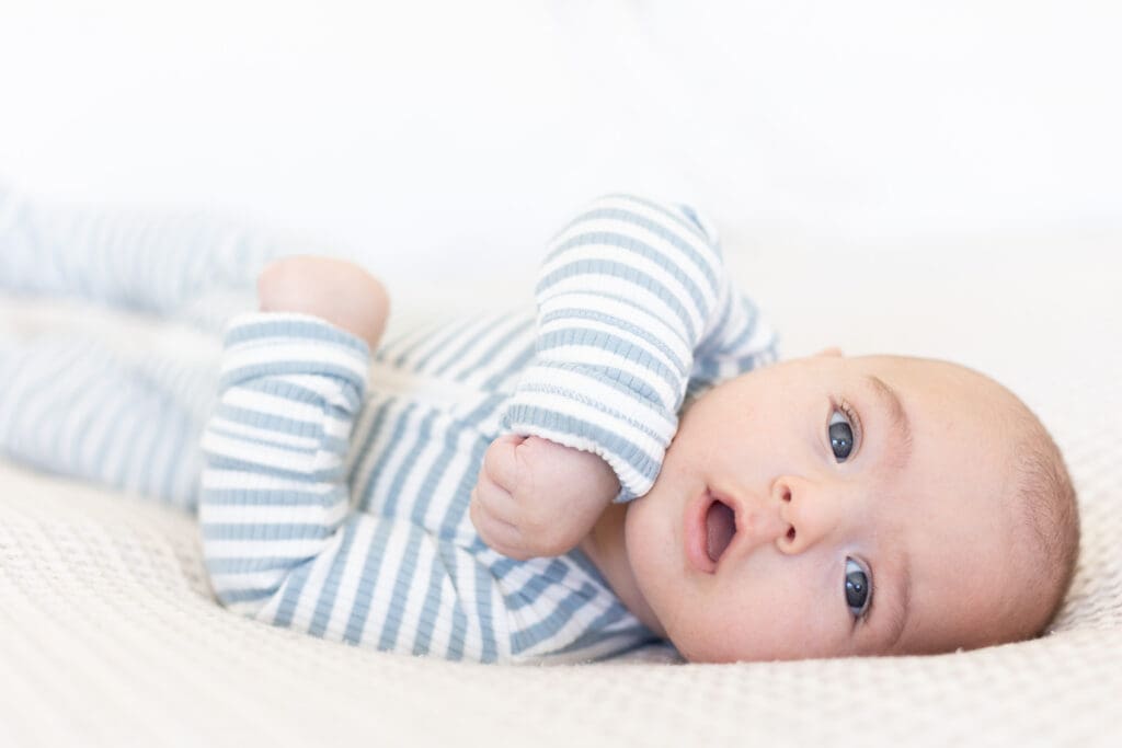 Close-up of a baby in a striped onesie lying on a soft cream blanket, looking curiously at the camera.