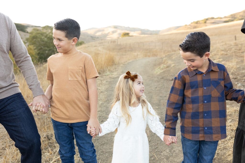 Three young siblings walking hand-in-hand with parents on a dirt trail through golden hills at sunset.