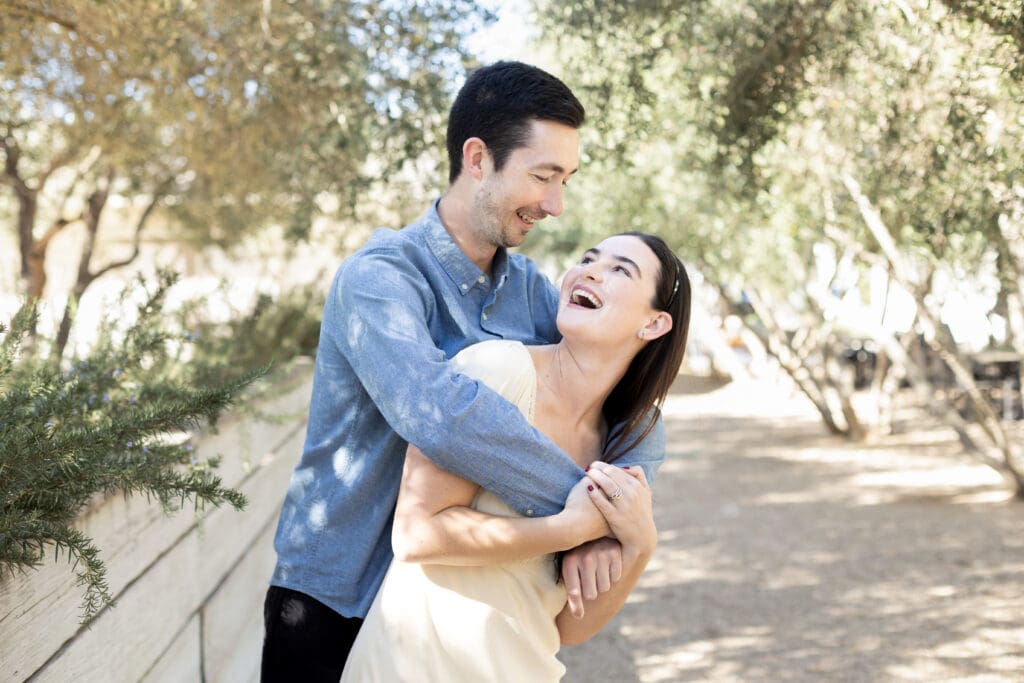 Couple laughing and embracing under dappled sunlight in a tree-lined outdoor path on a warm day.