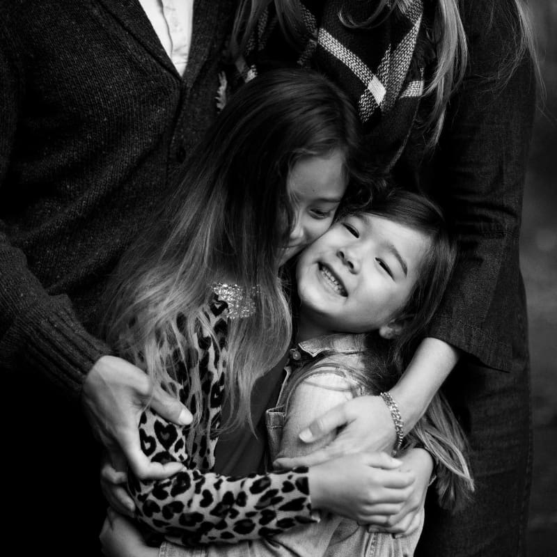 Two young sisters hugging and smiling during candid family moments, surrounded by their parents’ loving arms.