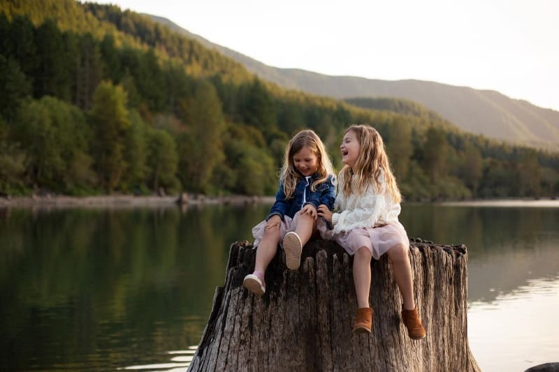Two young girls laughing on a tree stump by the lake, sharing joyful candid family moments outdoors.