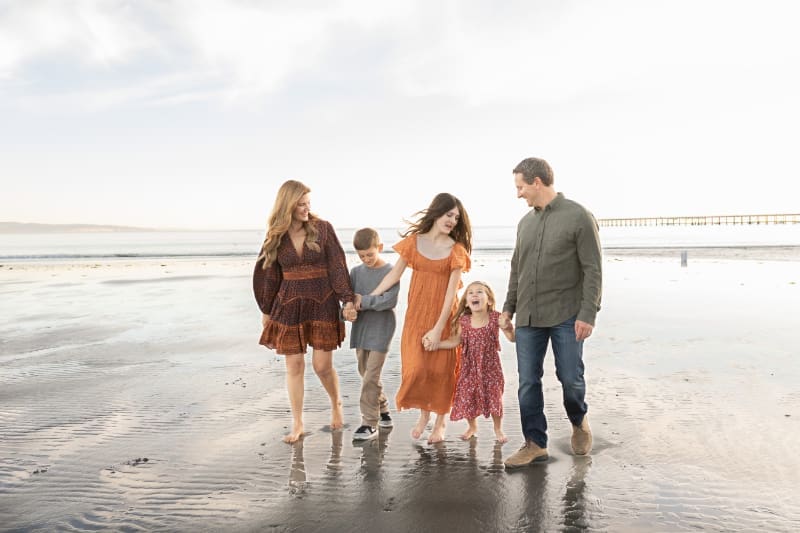 Family walking barefoot along the beach at sunset, sharing lighthearted candid family moments by the water.