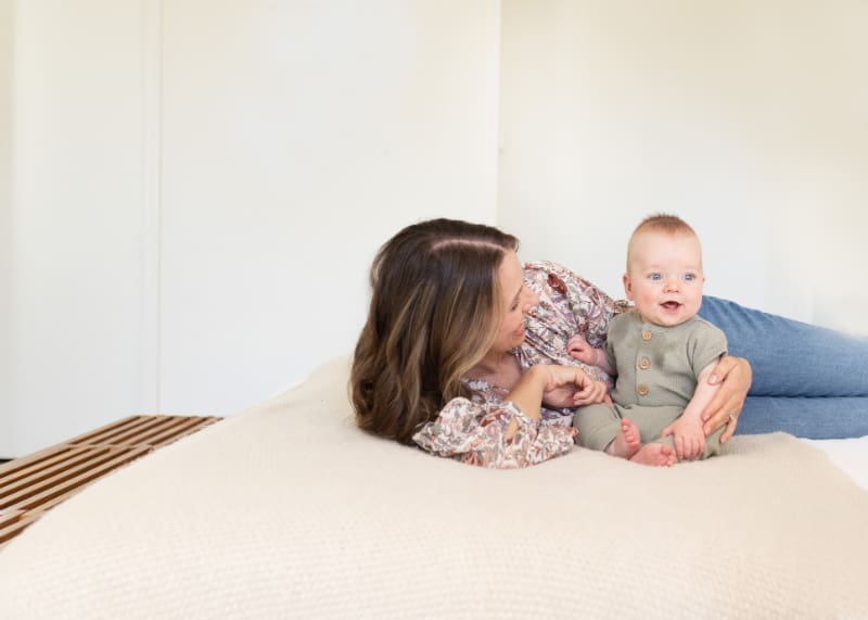 Mother lying on a bed with smiling baby in her lap, sharing a quiet, joyful moment together.