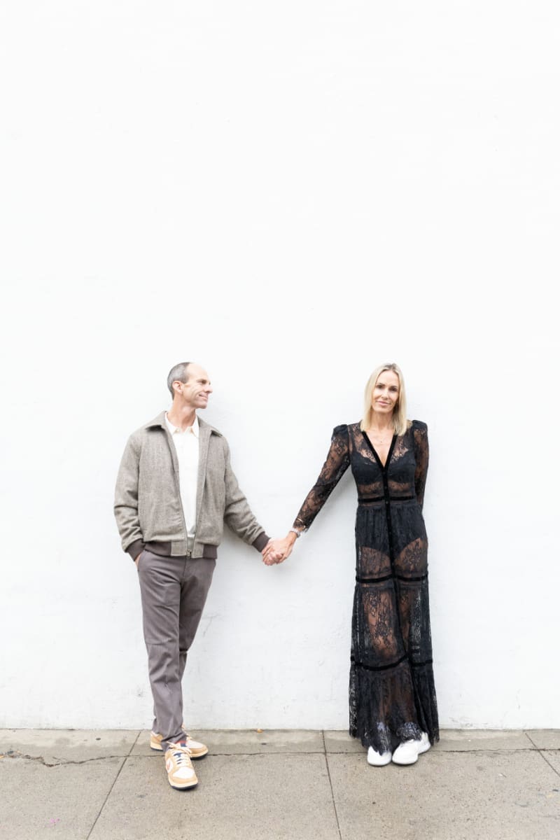 Couple holding hands and standing against a white wall, sharing a calm and stylish moment together.