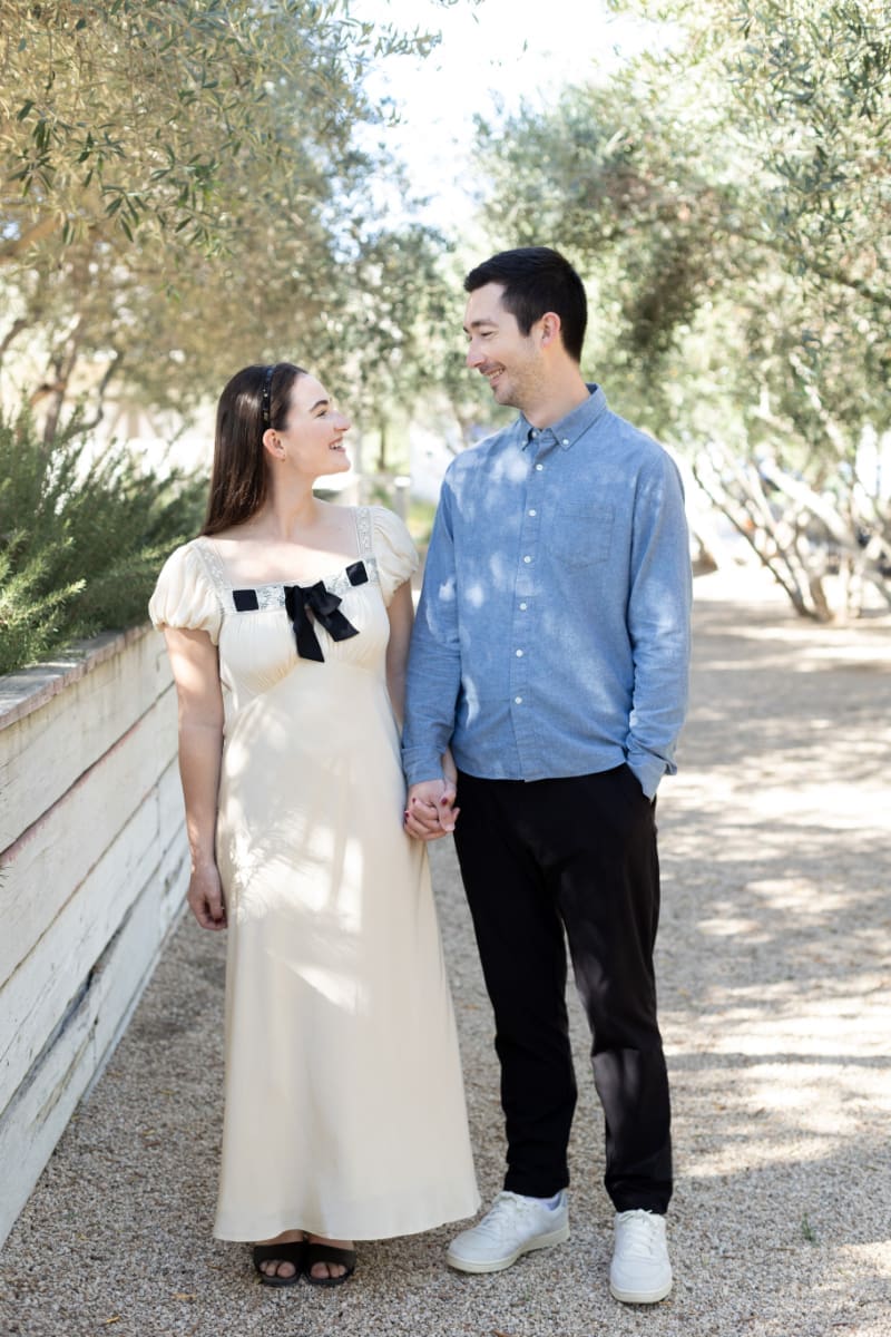 Couple holding hands and smiling at each other while walking outdoors on a sunlit path.
