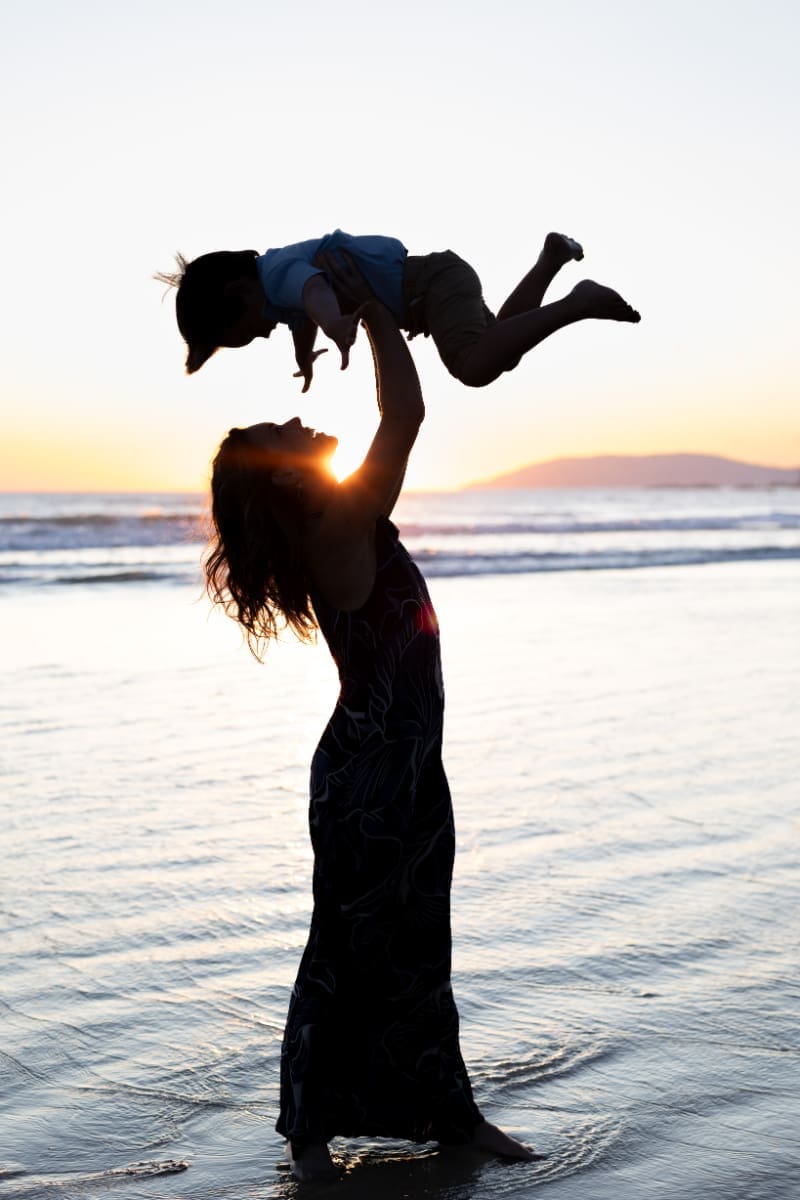 Silhouette of mother lifting child into the air at sunset on the beach, glowing with joy and light.