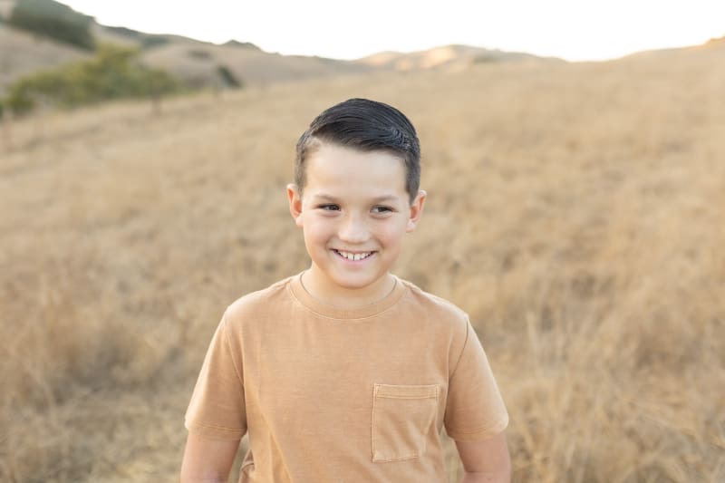 Smiling boy stands in a golden field during sunset, surrounded by soft hills and warm light.