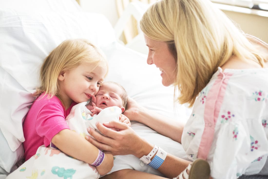 Candid newborn family photos of big sister kissing newborn in hospital bed