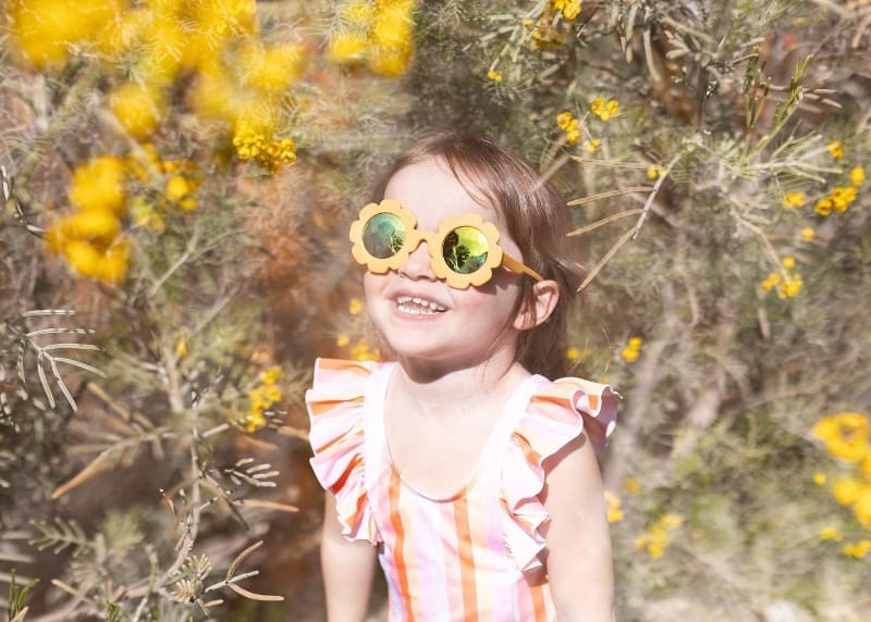 Smiling young girl wearing flower sunglasses in bright yellow wildflowers outdoors.