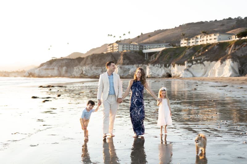 Beach family photos of parents and two kids walking barefoot together along calm shoreline at sunset