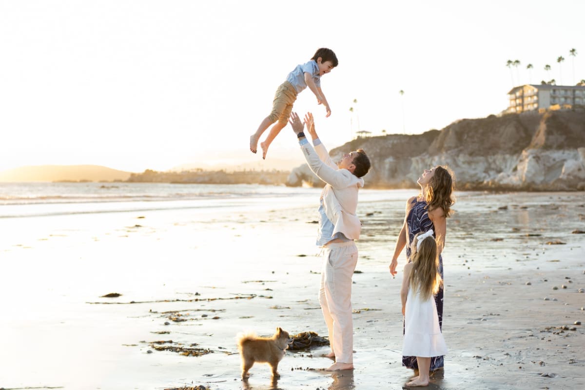 Father lifting young child into the air while family watches and smiles on beach at sunset
