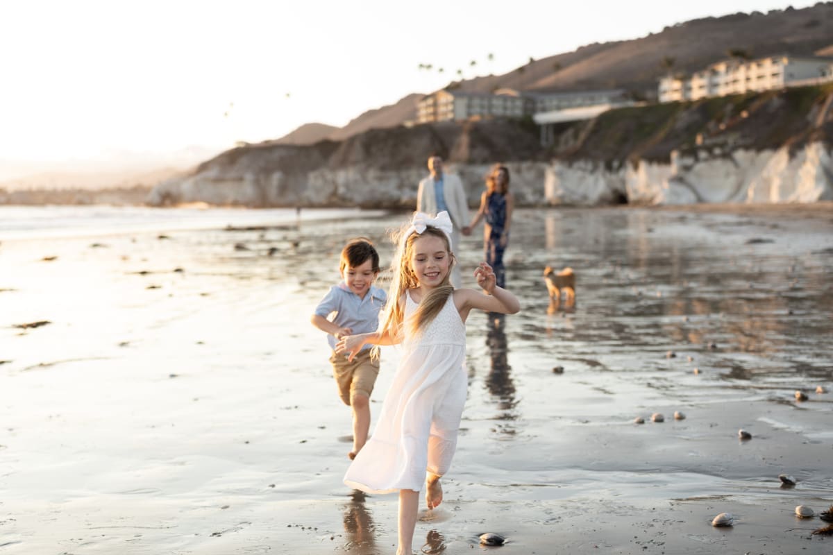 Beach family photos of kids running and laughing toward camera while parents walk behind near ocean shoreline