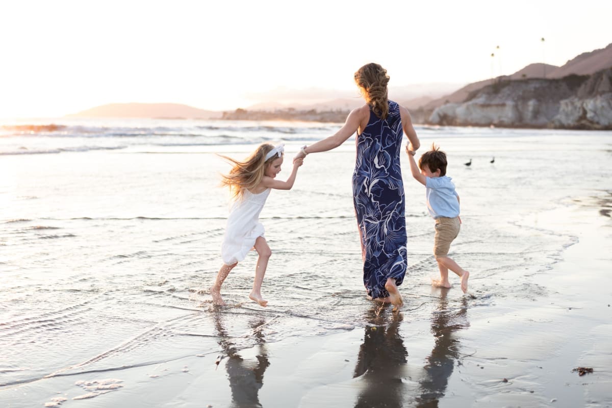 Mother holding hands with two children walking through shallow waves at sunset along the beach
