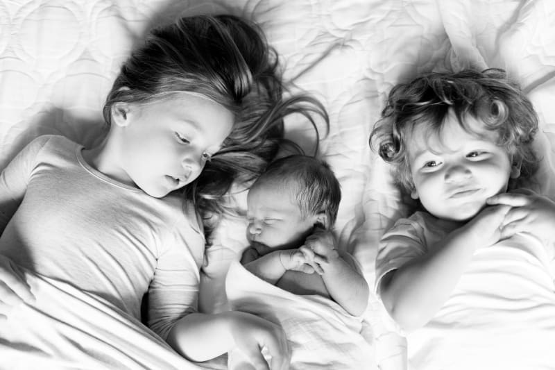 Older siblings lying beside their newborn baby on a bed, sharing a quiet moment together.