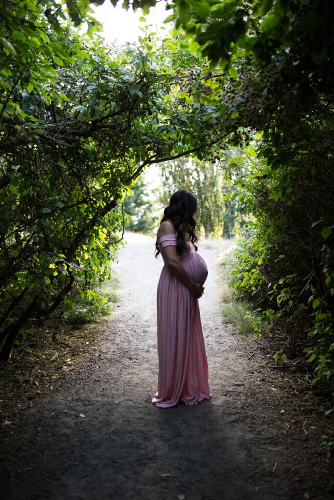 Pregnant mother in a flowing pink dress standing on a shaded path surrounded by lush green foliage during an outdoor maternity session.