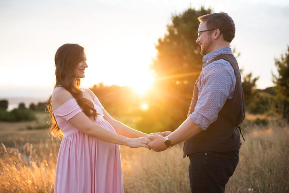 Pregnant mother and partner holding hands in a sunlit field during a golden hour maternity session.