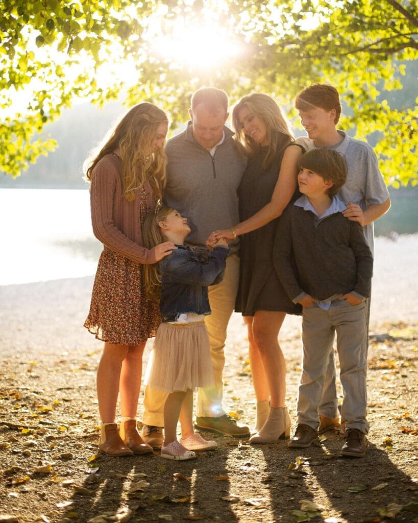 Family of six sharing a warm moment together beneath sunlit trees beside the water.